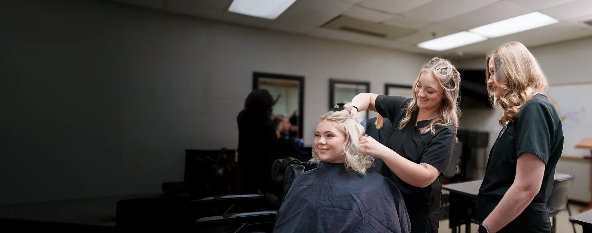 Student styles a client&rsquo;s hair while another student observes in a cosmetology classroom salon.