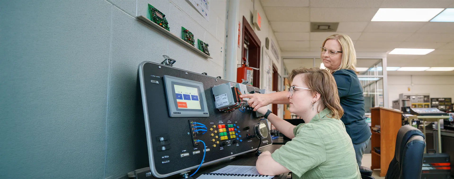 Instructor points to wiring on an industrial control training panel while a student operates the equipment in a lab.