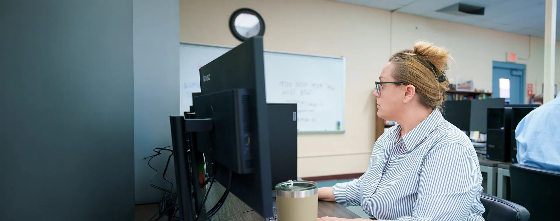 Person works at a desktop computer in a graphic design classroom with multiple workstations and a whiteboard.