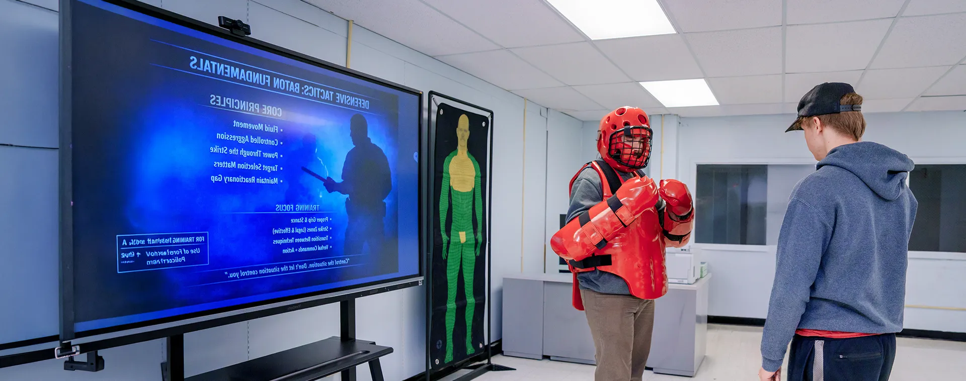 Person wearing red protective training gear stands in a classroom facing another person beside a large display screen.