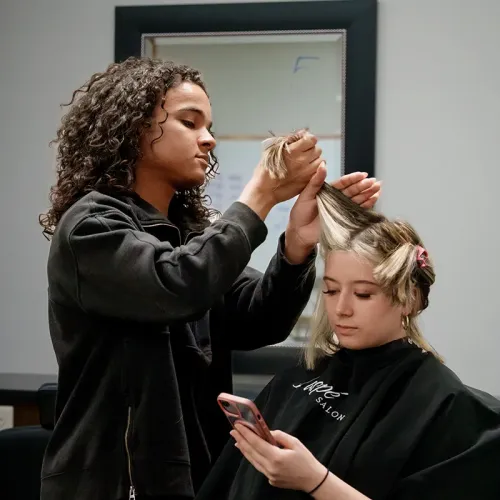 Student sections and styles hair for a seated client in a cosmetology training salon.