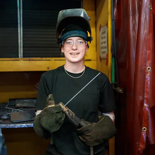 Welding student wearing a raised helmet, safety glasses, and gloves holds a welding electrode in a shop.