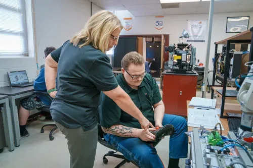 Instructor points to a handheld controller while assisting a student beside industrial automation equipment.