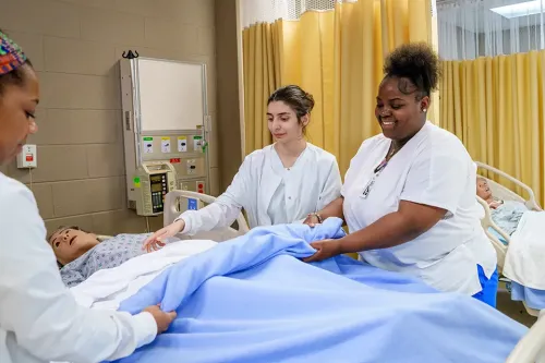Students in clinical uniforms make a bed around a patient care mannequin in a nursing skills lab.