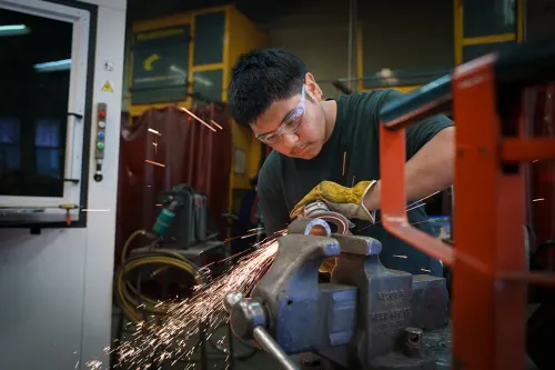 Student wearing safety glasses uses a grinder on metal secured in a bench vise, creating sparks.